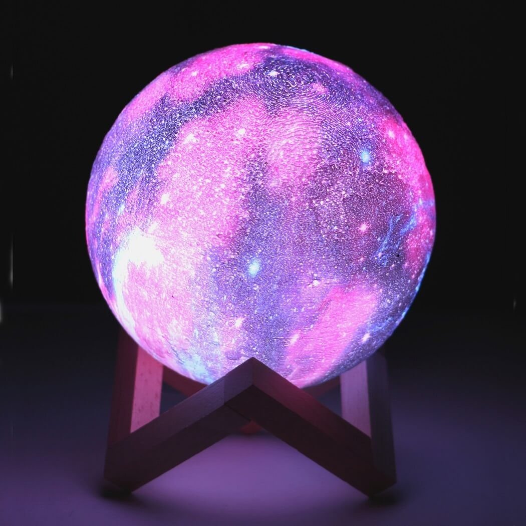 Lampe veilleuse galaxie violette et rose posée sur un support en bois, dans l’obscurité.