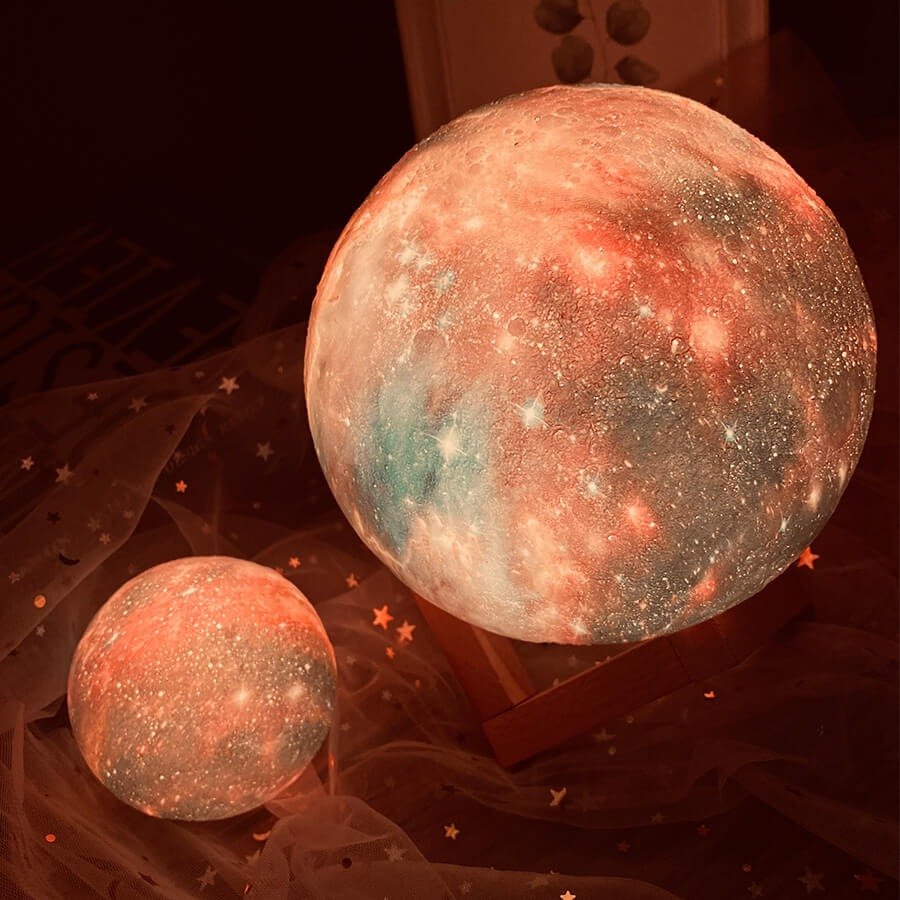 Deux lampes galaxie lune de tailles différentes avec des tons orangés et corail sur un tissu décoratif étoilé.