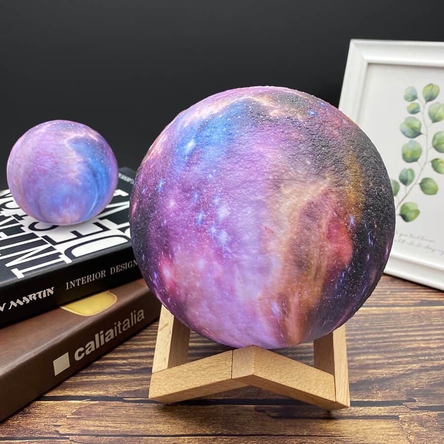 Lampe galaxie sphérique aux teintes violettes, roses et dorées, posée sur un support en bois; un second globe plus petit repose sur des livres à l’arrière-plan, devant un cadre botanique.
