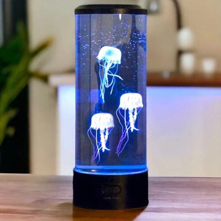 Lampe aquarium méduses cylindrique sur une table, avec trois méduses lumineuses blanches et bleues flottant dans un tube transparent, arrière‑plan flou