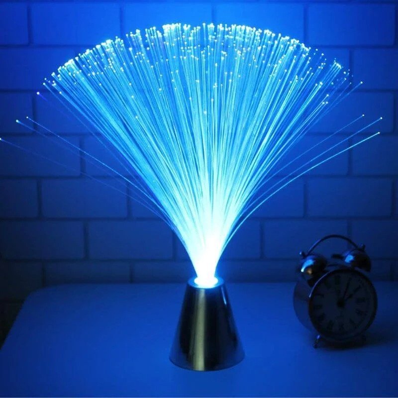 Lampe à fibre optique bleue avec faisceaux lumineux jaillissant d'une base conique, posée sur une table avec un réveil analogique en arrière-plan.