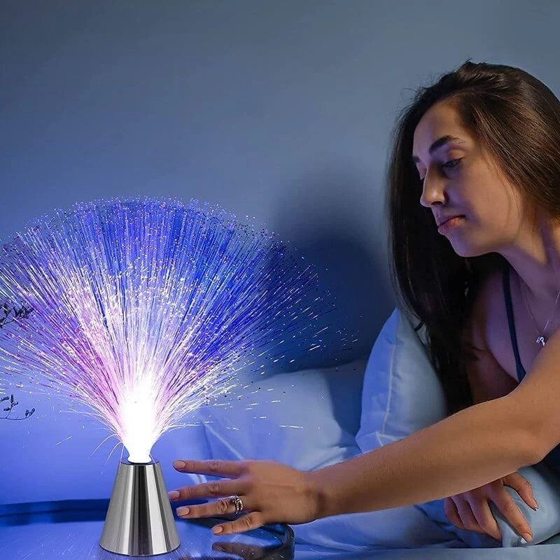 Lampe à fibre optique multicolore en faisceaux de lumière sur une base conique, avec une femme qui tend la main pour toucher les fibres illuminées.