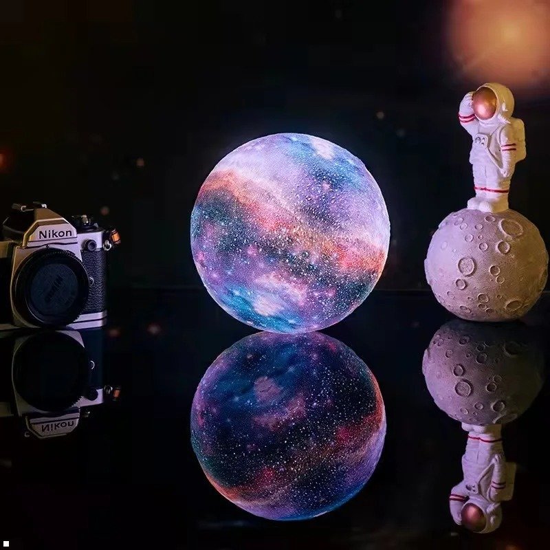 Lampe galaxie sphérique posée sur une surface réfléchissante, reflétant ses couleurs multicolores; une petite figurine d’astronaute debout sur un socle lunaire se tient à côté, avec un appareil photo Nikon en arrière-plan.