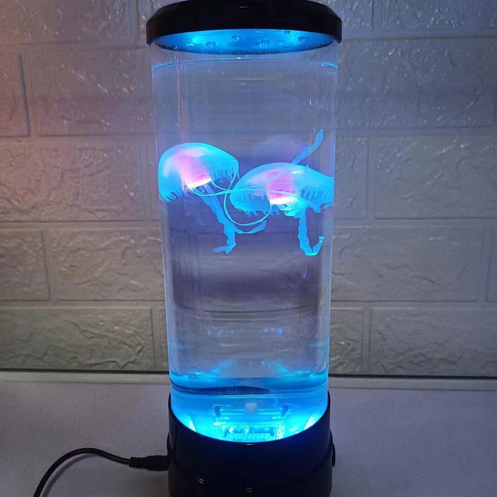 Lampe aquarium cylindrique avec deux méduses néon roses et bleues dans de l'eau claire, posée sur une surface blanche devant un mur en briques